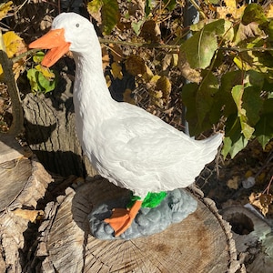 May include: White goose garden statue with orange beak and green feet. The goose is standing on a gray rock base and is positioned on a tree stump.