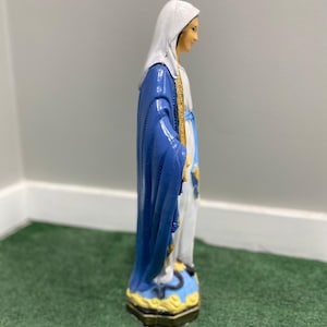 May include: A blue and white figurine of the Virgin Mary, standing on a yellow and blue base. The figurine is made of resin and is painted in a realistic style.
