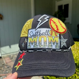 Puede incluir: Gorra de camionero negra de malla con una variedad de parches, incluyendo una pelota de softball, un rayo, un diamante y un parche que dice "Softball Mom" con un fondo de estampado de leopardo. La gorra tiene una cadena plateada alrededor del borde.