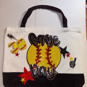 Może przedstawiać: Biała torba typu tote z czarnym dnem i uchwytami, z dużym cekinowym wzorem baseballowym z napisem "Game Day". Zawiera żółty i czerwony numer 38, gwiazdki i gest dłoni nr 1.