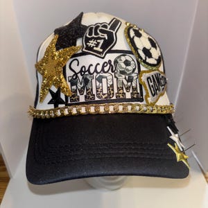 May include: A black and white trucker hat with a gold chain trim. The hat has several patches, including a gold star, a soccer ball, a hand with the number one, and a patch that says "Soccer Mom".