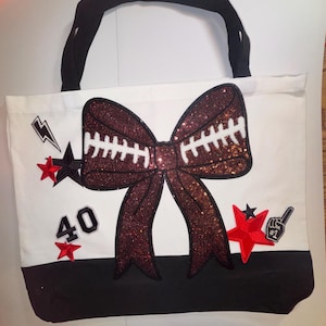 Może przedstawiać: Biała torba typu tote z dużą, cekinową kokardą w kształcie piłki nożnej. Torba ma czarne uchwyty i czarną podstawę. Ozdoby obejmują czerwone gwiazdy, błyskawicę, liczbę 40 i piankowy palec.