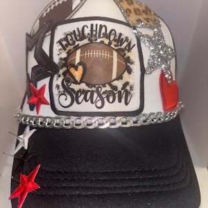 Könnte beinhalten: Weiße Trucker-Kappe mit schwarzem Schirm. Die Kappe hat verschiedene Patches, darunter ein Football, ein Herz, Sterne und ein Patch mit der Aufschrift "Touchdown Season".