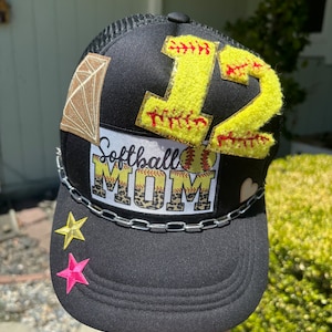 May include: Black mesh trucker hat with a chain detail. The hat has a variety of patches including a diamond, a softball with the number 12, and a patch that says "Softball Mom" with a leopard print background.