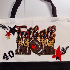 Może przedstawiać: Biała torba typu tote z czarnymi uchwytami i czarną podstawą. Torba zawiera słowa "Football Mom" w błyszczącej czerwieni i złocie, piłkę nożną z sercem oraz akcenty gwiazd i błyskawic. Zawiera również numer 40.