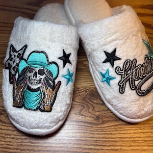 Könnte beinhalten: Weiße Plüschhausschuhe mit gestickten Designs. Ein Hausschuh zeigt einen Totenkopf mit türkisfarbenem Cowboyhut, mit dem Text "Howdy" auf dem anderen Hausschuh. Akzentuiert mit schwarzen und türkisfarbenen Sternen.