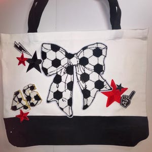 Może przedstawiać: Biała torba typu tote z czarnymi uchwytami i czarnym dołem. Torba posiada dużą kokardę z wzorami piłek nożnych, czerwone gwiazdy i numer 46. Widoczna jest również czarna naszywka z piankowym palcem nr 1.