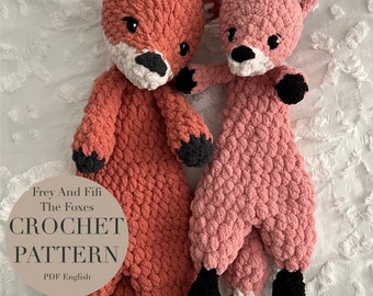 May include: Two crocheted fox plushies, one red and one pink, with black eyes and noses. The red fox is larger and has white accents on its paws. The pink fox is smaller and has a white accent on its tail. The text "Frey And Fifi The Foxes CROCHET PATTERN PDF English" is on a white circle.
