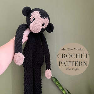 May include: A crocheted monkey toy with black fur and pink hands and feet. The monkey is being held by a hand. The text "Mel The Monkey CROCHET PATTERN PDF English" is on a brown circle in the background.