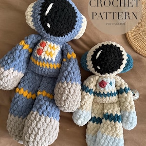 May include: Two crocheted astronaut dolls in blue, gray, and cream colors. The larger doll is blue with yellow accents, and the smaller doll is cream with blue stripes. The text "Cosmo&Cadet the Astronauts CROCHET PATTERN PDF ENGLISH" is visible.