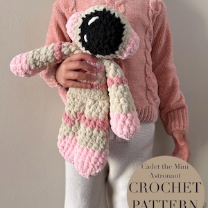 Könnte beinhalten: Handgefertigte Häkel-Astronautenpuppe in Creme, Rosa und Schwarz. Die Puppe hat ein großes schwarz-weißes Gesicht, rosa Akzente und wird von einer Person gehalten, die einen rosa Pullover trägt. Auf der Puppe steht "Cadet the Mini Astronaut CROCHET PATTERN PDF ENGLISH."