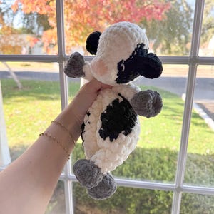 May include: A crocheted stuffed cow toy with black and white spots and gray hooves. The toy is being held by a hand in front of a window with fall foliage.