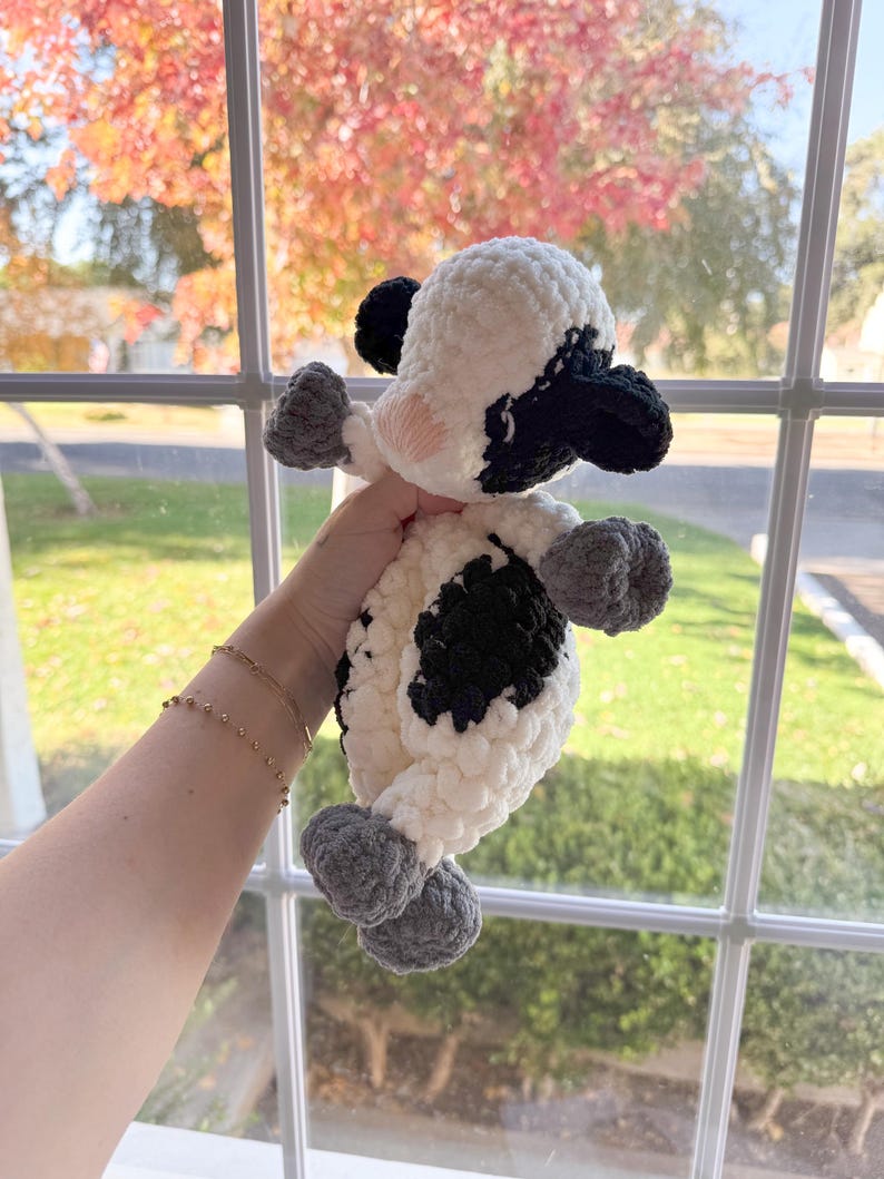 May include: A crocheted stuffed cow toy with black and white spots and gray hooves. The toy is being held by a hand in front of a window with fall foliage.