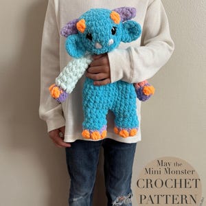 May include: A crocheted blue monster toy with orange, purple and white accents. The toy is being held by a person wearing blue jeans and a white long-sleeved shirt. The text "May the Mini Monster CROCHET PATTERN PDF English" is visible in the image.