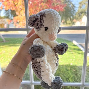 May include: A crocheted stuffed animal shaped like a calf. The calf is white with gray spots and has a pink nose. The calf is being held by a hand.