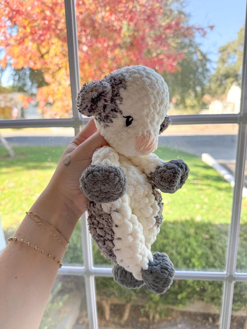 May include: A crocheted stuffed animal shaped like a calf. The calf is white with gray spots and has a pink nose. The calf is being held by a hand.