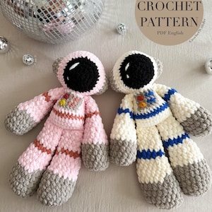 以下が含まれることがあります： 手作りの宇宙飛行士かぎ針編み人形が2体。1つは茶色のストライプが入ったピンク色、もう1つは青色のストライプが入った白色です。どちらも黒いバイザーとグレーのアクセントがあり、明るい表面に置かれています。「Cosmo Astronaut CROCHET PATTERN PDF English」というテキストが含まれています。