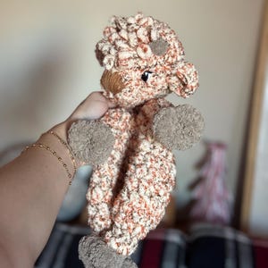 May include: A crocheted stuffed animal in the shape of a brown and white sheep. The sheep has a brown nose and black eyes. The sheep is being held by a hand.