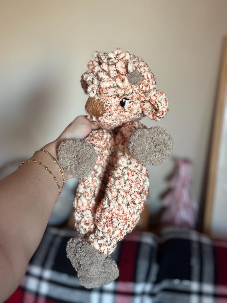 May include: A crocheted stuffed animal in the shape of a brown and white sheep. The sheep has a brown nose and black eyes. The sheep is being held by a hand.