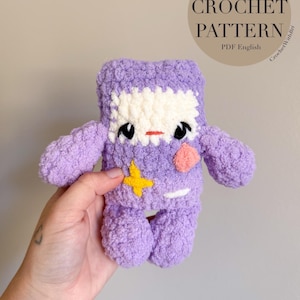 以下が含まれることがあります： 手作りの紫色の編み人形。白い顔、黒い目、小さな赤い口があります。人形の体には黄色の星とピンクのハートが付いています。画像には「Yarn Boy Crochet Pattern PDF English」というテキストも含まれています。