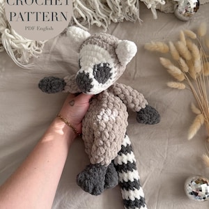 May include: A crocheted lemur toy with a gray and white striped tail. The lemur is made with a soft, fluffy yarn and has a gray face with black eyes. The toy is being held by a hand.