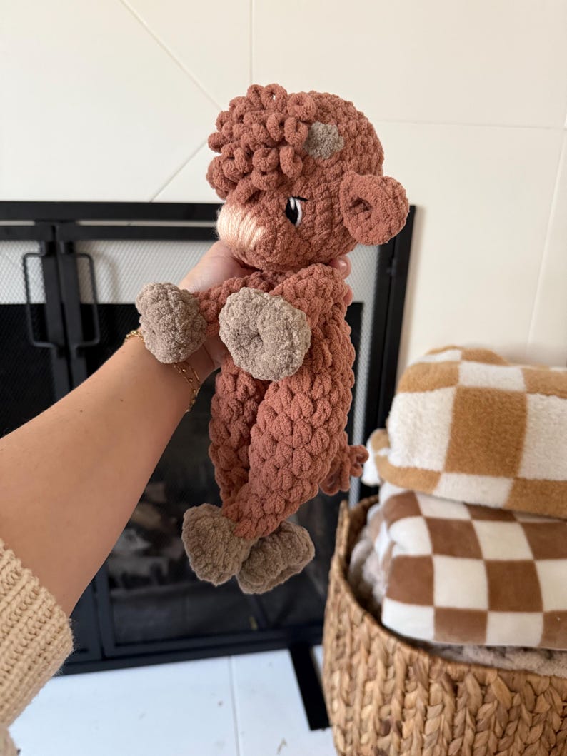 May include: A brown crocheted stuffed animal shaped like a cow with a white spot on its head. The cow is being held by a hand.