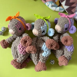 May include: Three crocheted plush toys in different colors. The toys are brown with pink, blue, and purple accents. Each toy has a different colored ribbon tied around its head. The toys are sitting on a green surface.