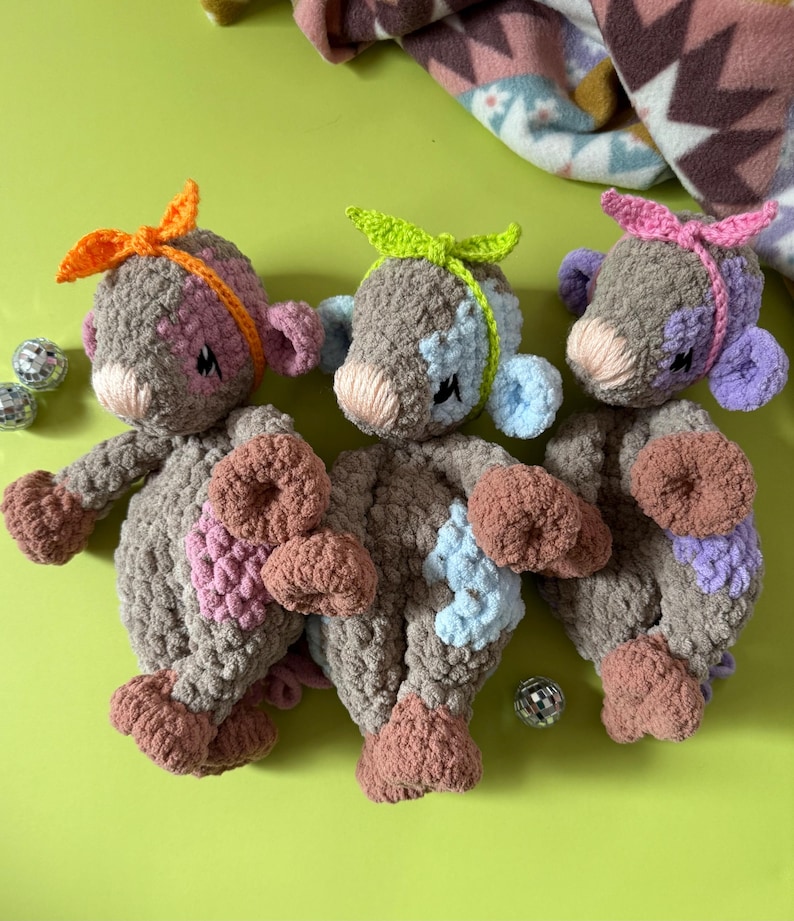 May include: Three crocheted plush toys in different colors. The toys are brown with pink, blue, and purple accents. Each toy has a different colored ribbon tied around its head. The toys are sitting on a green surface.