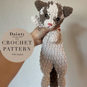 以下が含まれることがあります： 茶色と白のぬいぐるみ猫の編み物パターン。パターン名は「Dainty」で、英語のPDFで入手できます。