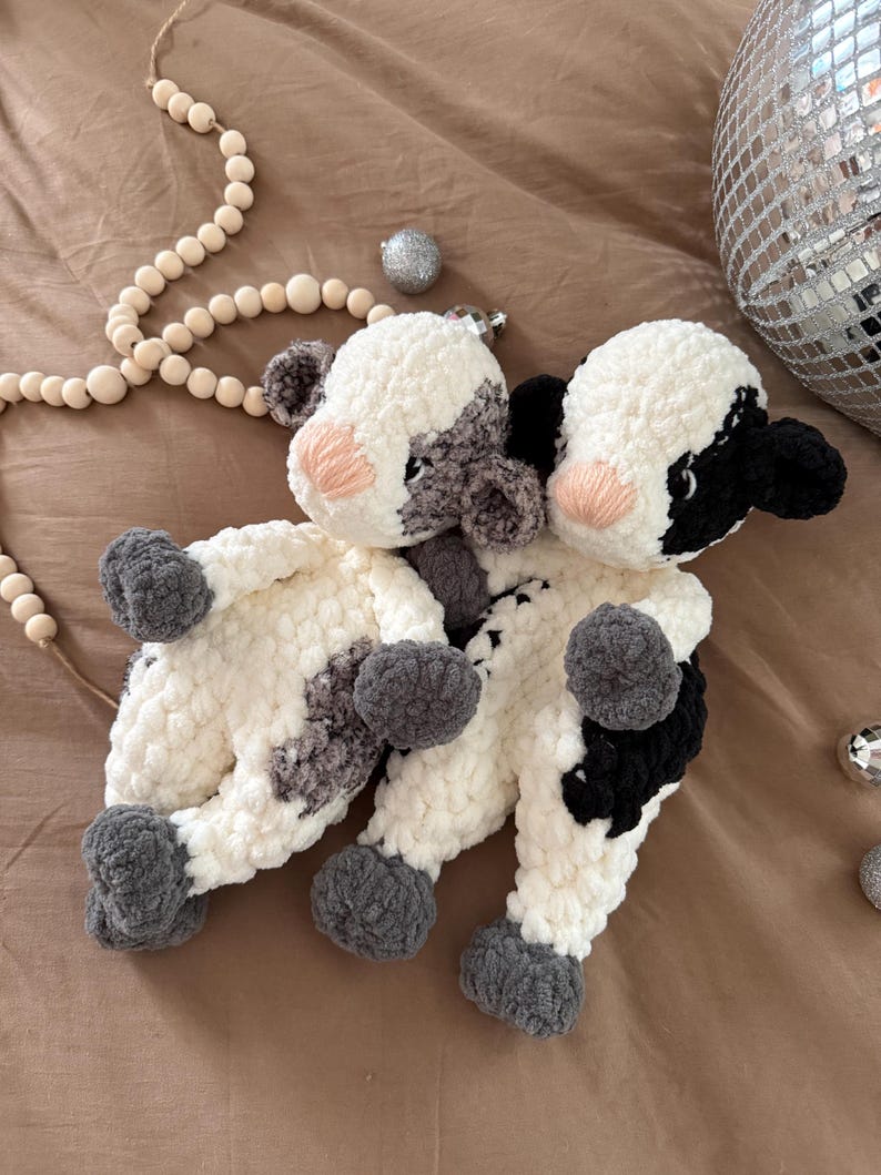 May include: Two crocheted plush cow toys with black and white patches. The cows are sitting on a brown surface.