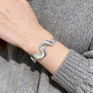 May include: A polished silver cuff bracelet featuring a continuous, fluid wave-like design. The metal has a smooth, reflective surface that catches the light, creating a series of gentle curves that wrap around the wrist in a singular, solid band.