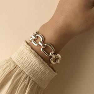 May include: A polished silver-tone chain link bracelet featuring thick, rounded rectangular links connected in a repeating pattern. The bracelet is worn around the wrist, showcasing a smooth, reflective metallic surface and a sturdy construction.