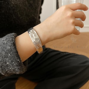 May include: A silver-toned metal cuff bracelet with a hammered, textured surface. The bracelet features a wide, slightly curved band design that wraps around the wrist, showcasing a reflective finish and irregular indentations across the exterior.