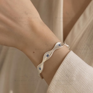 May include: A silver bangle bracelet with a wavy, textured surface featuring three small, dark blue circular gemstones set within star-shaped engravings. The metal has a brushed finish, and the design follows a continuous undulating pattern.