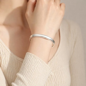 May include: A smooth, polished silver-toned bangle bracelet with a rounded, continuous band design. The bracelet features a clean, reflective surface and a simple, solid construction that encircles the wrist with a uniform, sleek appearance.