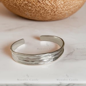May include: A silver-toned metal cuff bracelet featuring an open design and a textured, wavy surface with horizontal ridges. The bracelet has a polished finish and a curved, organic shape that tapers slightly at the ends for a comfortable fit.