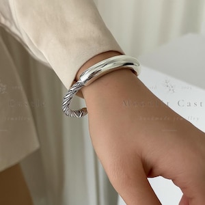 May include: A silver cuff bracelet with a rounded top and a twisted rope design on the bottom half. The bracelet is worn on a wrist, showcasing its polished, reflective surface. The jewelry is a simple, elegant accessory.