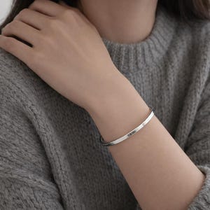 May include: A simple, polished silver-toned metal bangle bracelet with a smooth, rounded surface. The bracelet features a clean, minimalist design with a consistent thickness throughout, encircling the wrist with a reflective, metallic finish.
