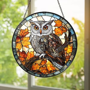 May include: A round stained glass window featuring an owl perched on a branch with autumn leaves. The owl has detailed feather patterns in shades of gray, brown, and white, with bright orange eyes. The leaves are in shades of orange and yellow.