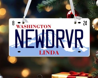 May include: A rectangular white hanging ornament shaped like a Washington license plate. It features the text NEWDRVR in large dark blue letters, the word WASHINGTON in red, and the name LINDA in red. A light blue mountain graphic is visible.