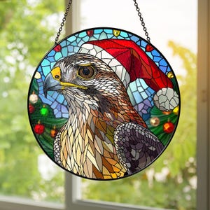 May include: Stained glass round ornament featuring a hawk wearing a Santa hat. The hawk has brown, tan, and white feathers, a yellow beak, and a dark eye. The background is blue with a Christmas tree and ornaments.