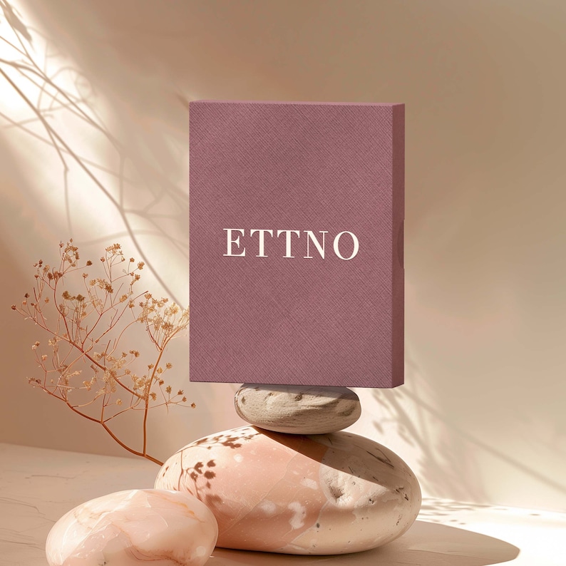 May include: A mauve box with the word "ETTNO" printed in white on the front, sitting on top of three stacked, light-colored stones.