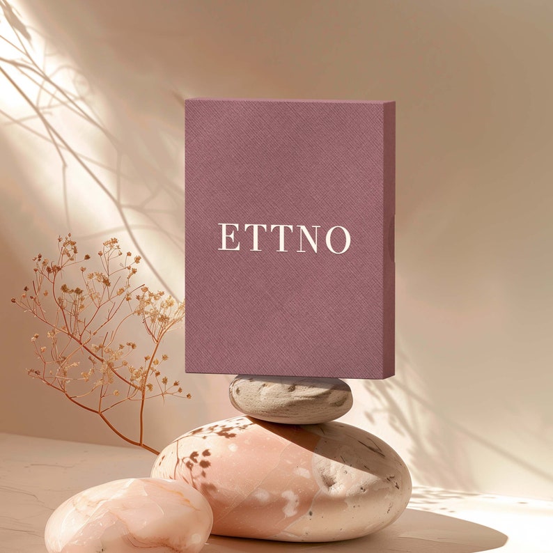 May include: A mauve box with the word "ETTNO" printed in white on the front, sitting on top of three stacked, light-colored stones.