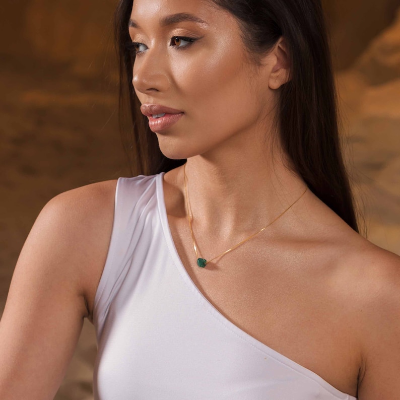 May include: A woman wearing a white one-shoulder top with a gold chain necklace featuring a dark green gemstone pendant.