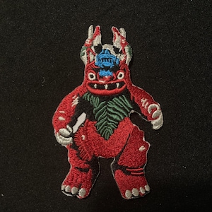 May include: Embroidered patch featuring a stylized red monster with a blue headpiece and green accents. The patch has a detailed design with silver and white highlights, and is likely for clothing or accessories.