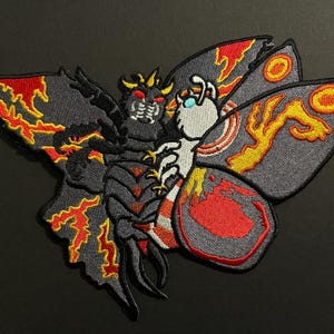 May include: Embroidered patch featuring a stylized moth-like creature with a detailed design. The patch showcases a dark gray body with red and yellow accents on the wings and body. The creature has a detailed face and a light blue gem.