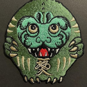 May include: Embroidered patch featuring a green monster with a detailed design. The monster has large eyes, sharp teeth, and a red mouth. The patch includes Japanese characters and gold-colored claws.