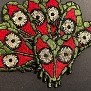 Puede incluir: Parches bordados con un diseño en forma de corazón con un centro rojo, borde verde y grandes detalles en forma de ojo con un centro plateado. Los parches están apilados, mostrando la intrincada costura y la paleta de colores.