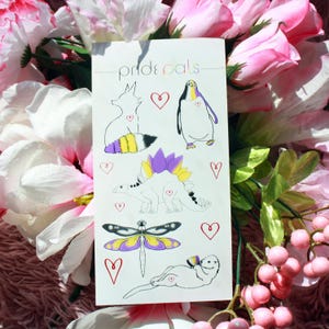 May include: A white card featuring the text "Pride Pals" in rainbow colors, with illustrations of a raccoon, penguin, stegosaurus, dragonfly, and otter, each with a heart. The raccoon's tail and the otter's head have non-binary flag colors.