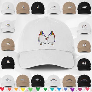 May include: A white baseball cap featuring an embroidered graphic of two penguins holding hands, each with a rainbow-coloured back and a small red heart on its chest. The image also displays a grid of various baseball caps in white, tan, and black.